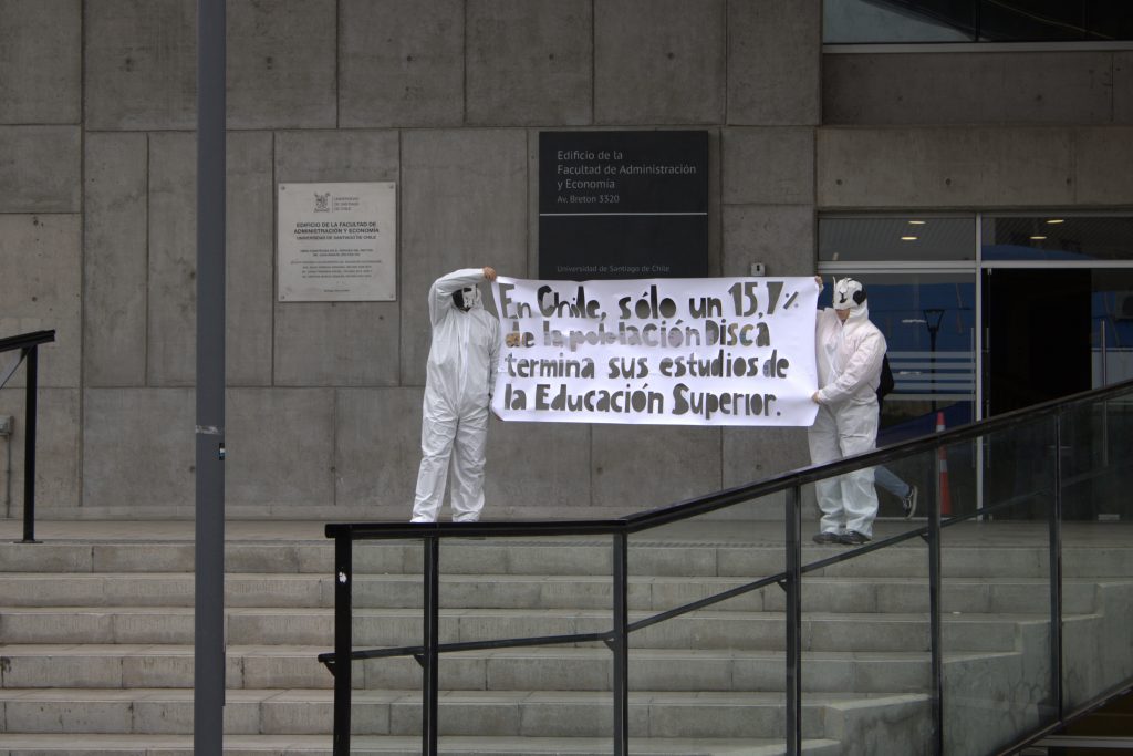 Dos personas en overoll blanco sosteniendo un carte que dice "En Chile, solo un 15% de la población disca termina sus estudios de la Educación Superior"